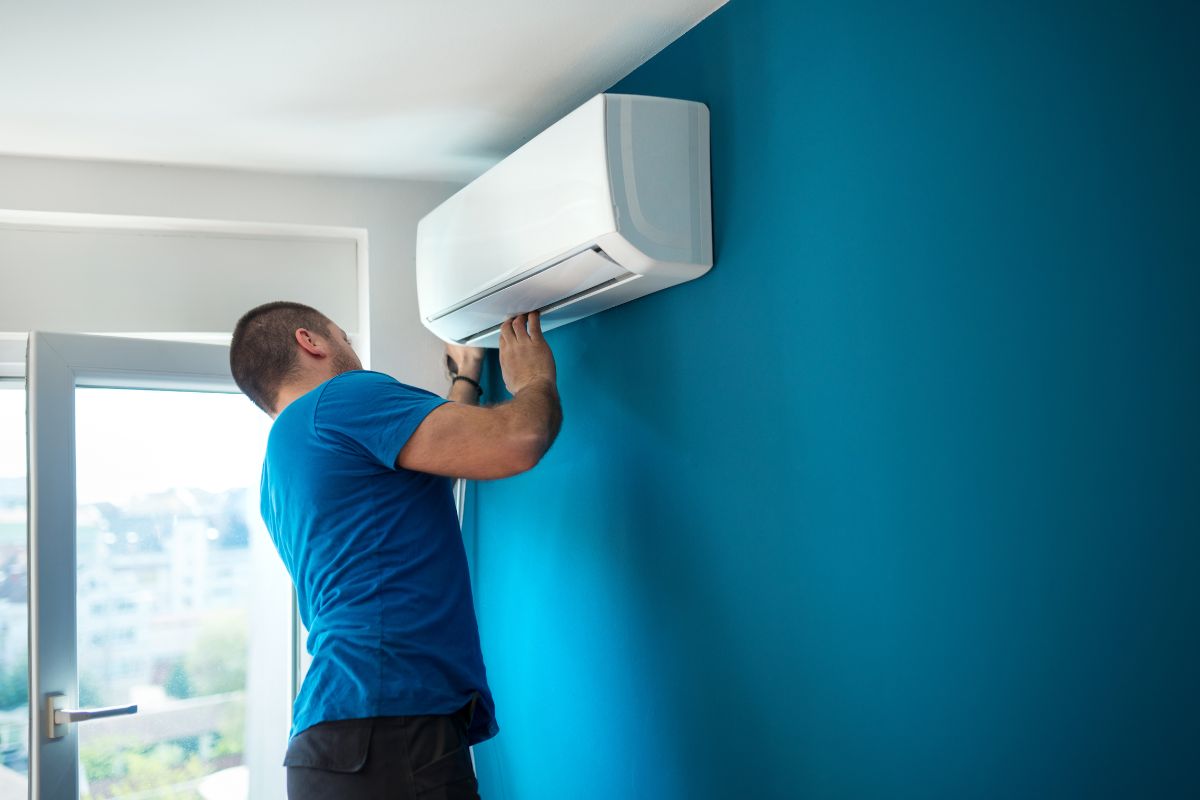 A man repairs an air conditioner in a room, using tools and focused on the unit's internal components. 