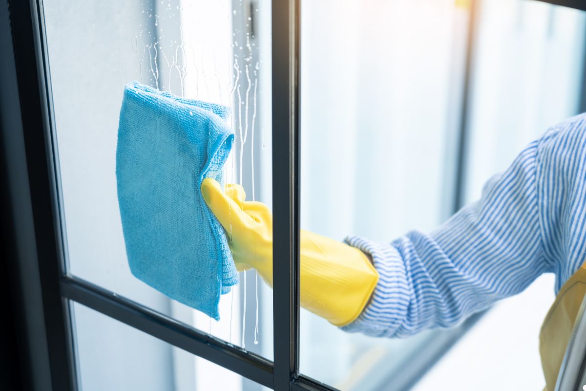 A person wearing an apron and rubber gloves is cleaning a window with a cloth, focusing on removing dirt and smudges.